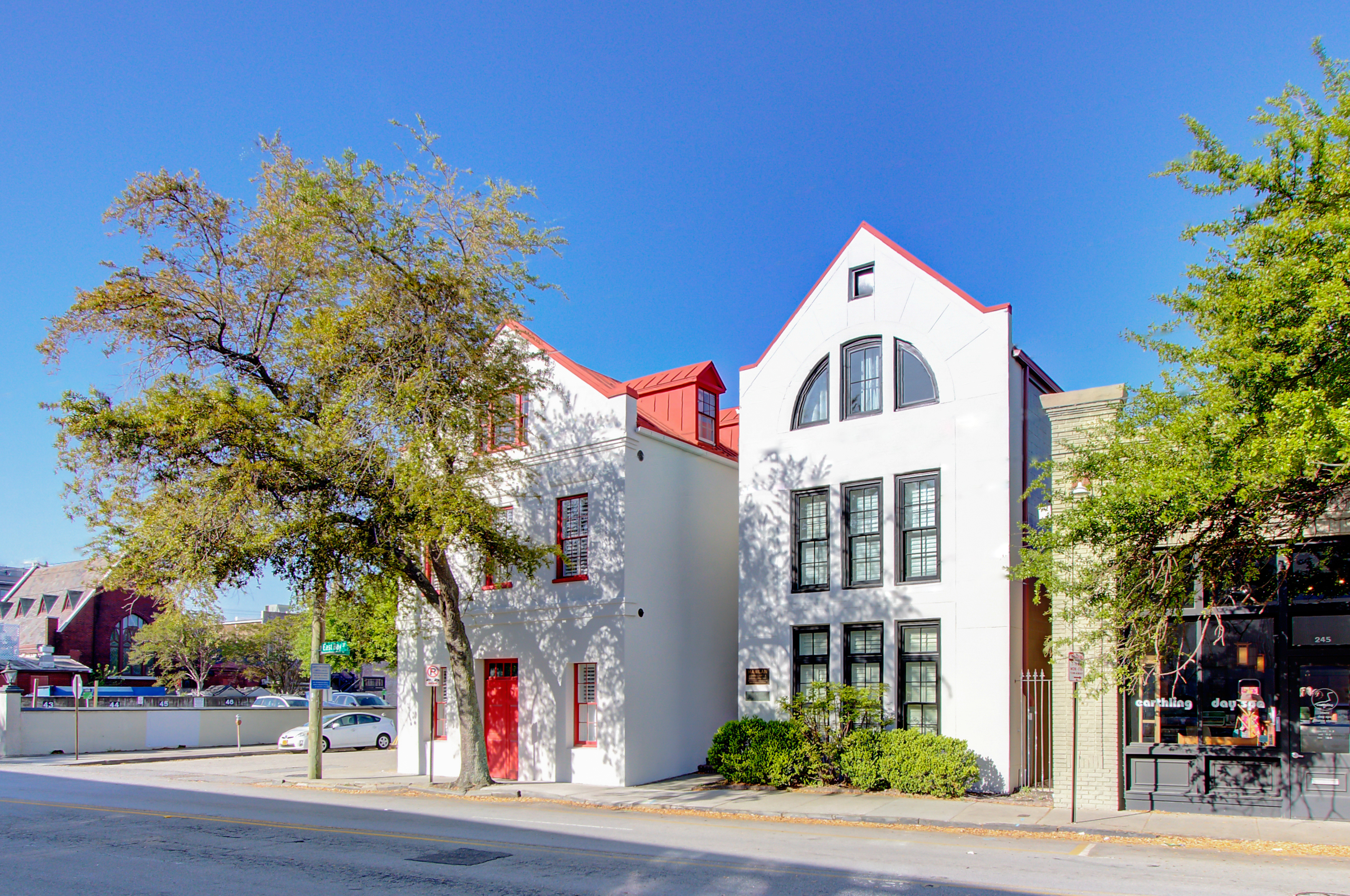 A white building with red trim and a red roof stands on a street corner with trees and a clear blue sky.